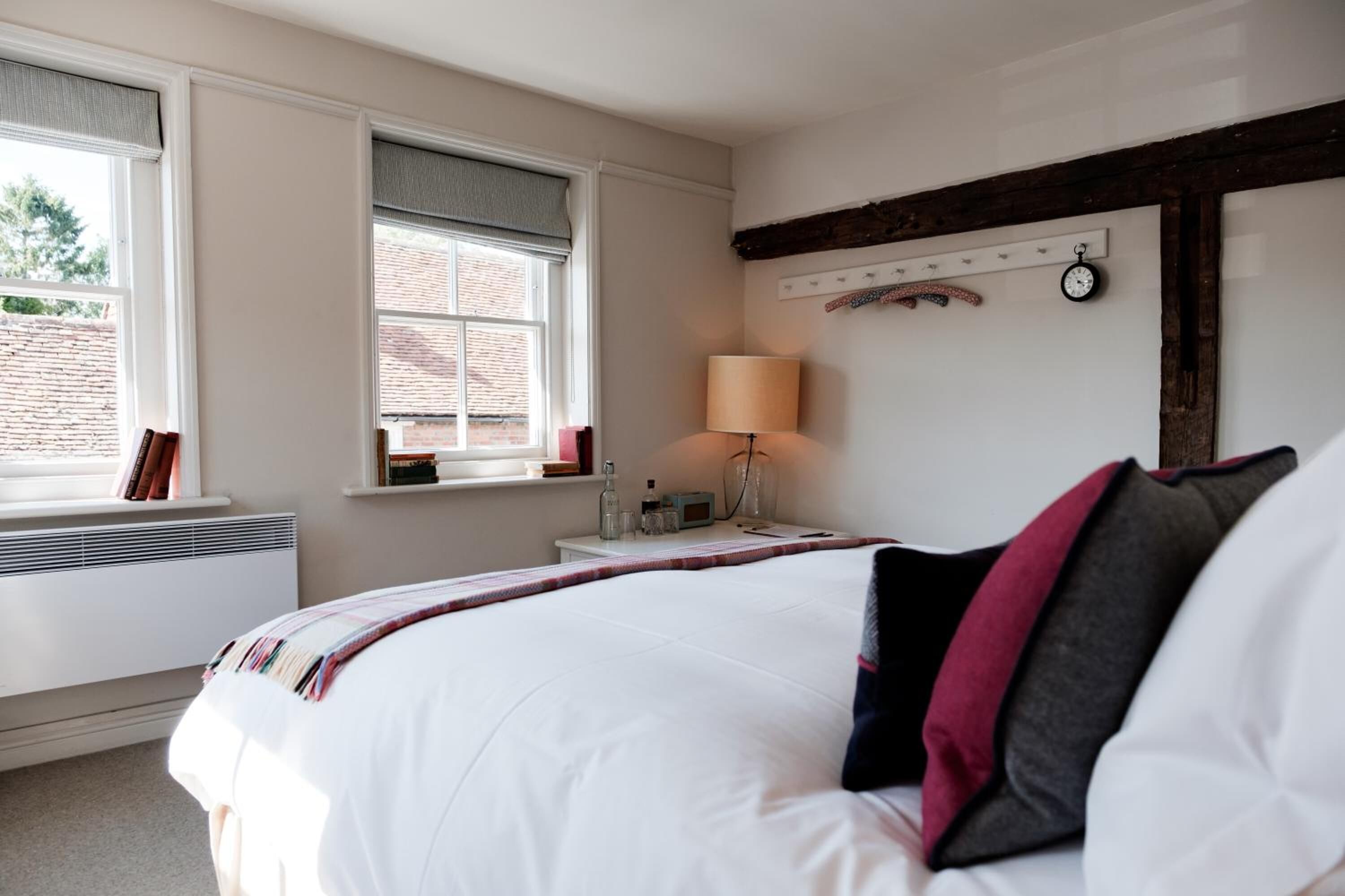 A bedroom with a large bed, a bedside table with a lamp, a window with blinds, and a wall-mounted clock.