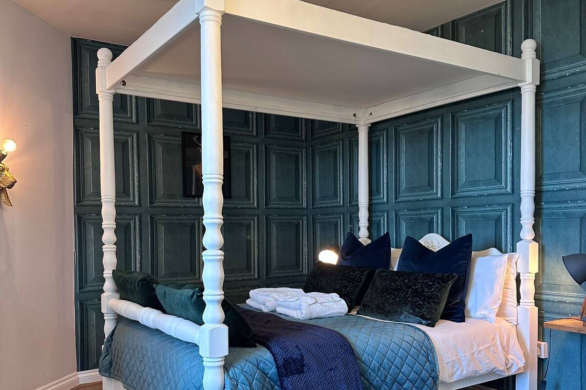Uma cama com dossel com edredom acolchoado azul e branco, um tapete azul escuro e um candelabro com lâmpadas aparentes.