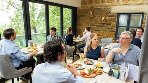 Un groupe de personnes est réuni dans un restaurant qui possède de grandes fenêtres, des murs en pierre et une table en bois.
