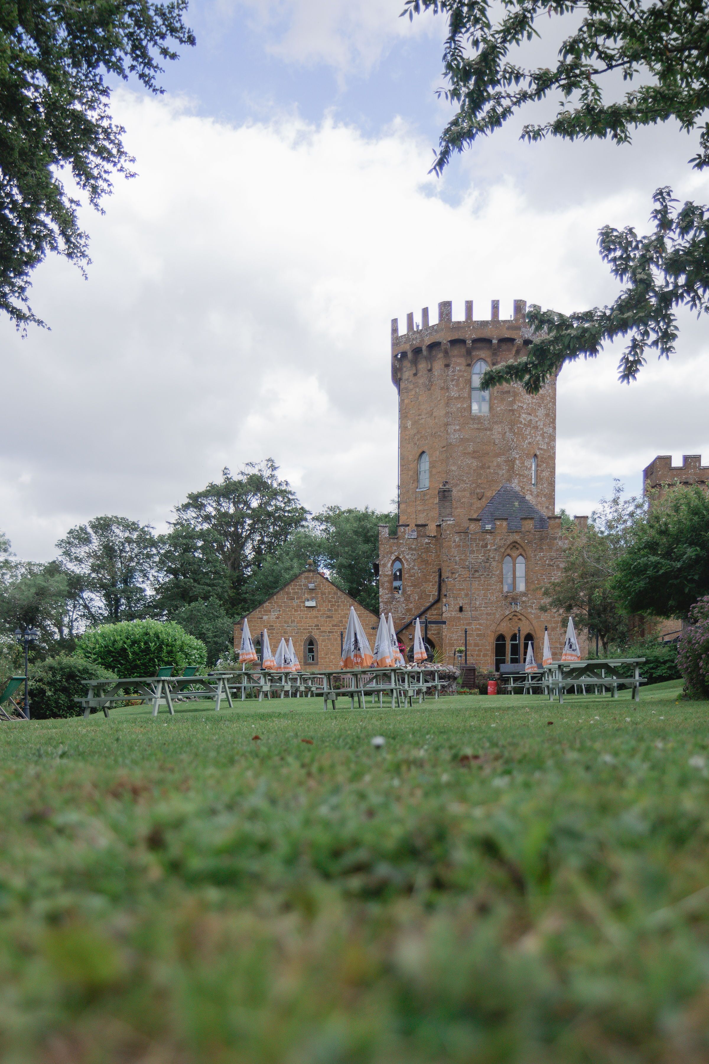 Een kasteel met een hoge toren, omgeven door groen en een gazon met picknicktafels en parasols.