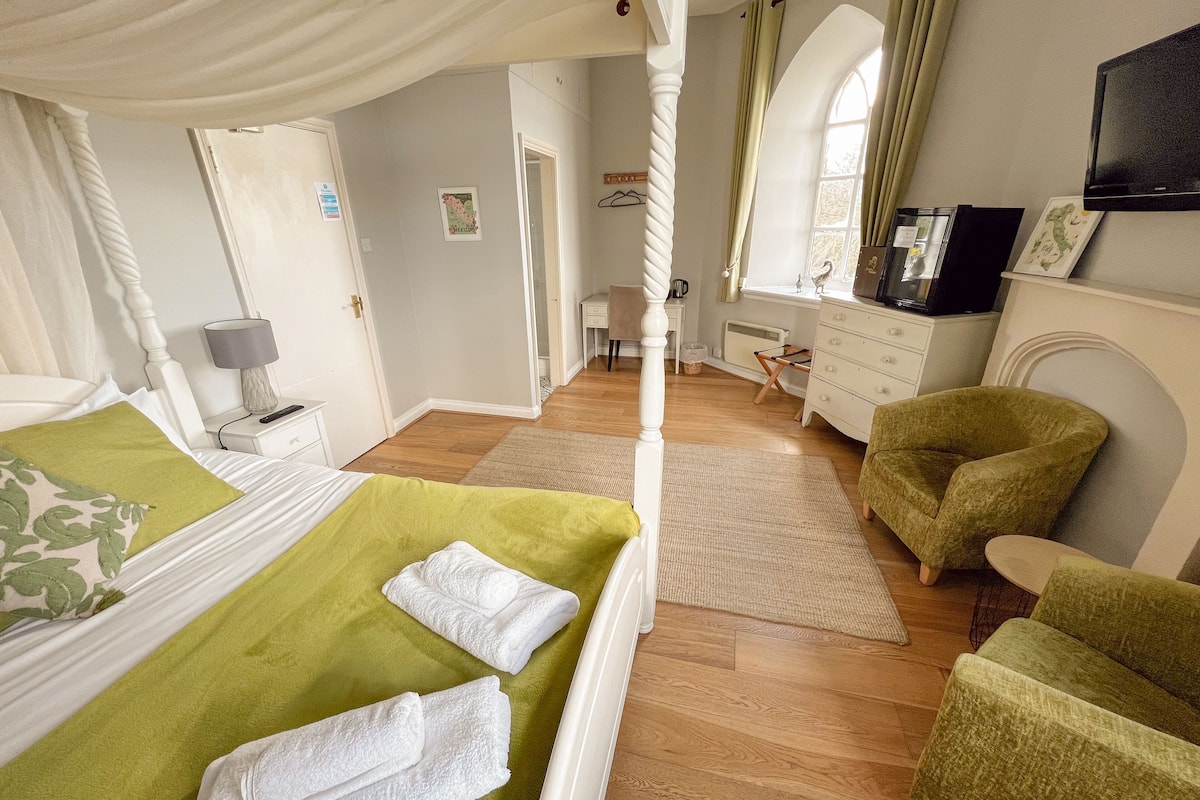 Quarto com cama de dossel, colcha verde e toalhas brancas. Há uma poltrona verde e um aparador de madeira.