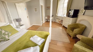 A bedroom with a four-poster bed, a green bedspread, and white towels. There is a green armchair and a wooden dresser.