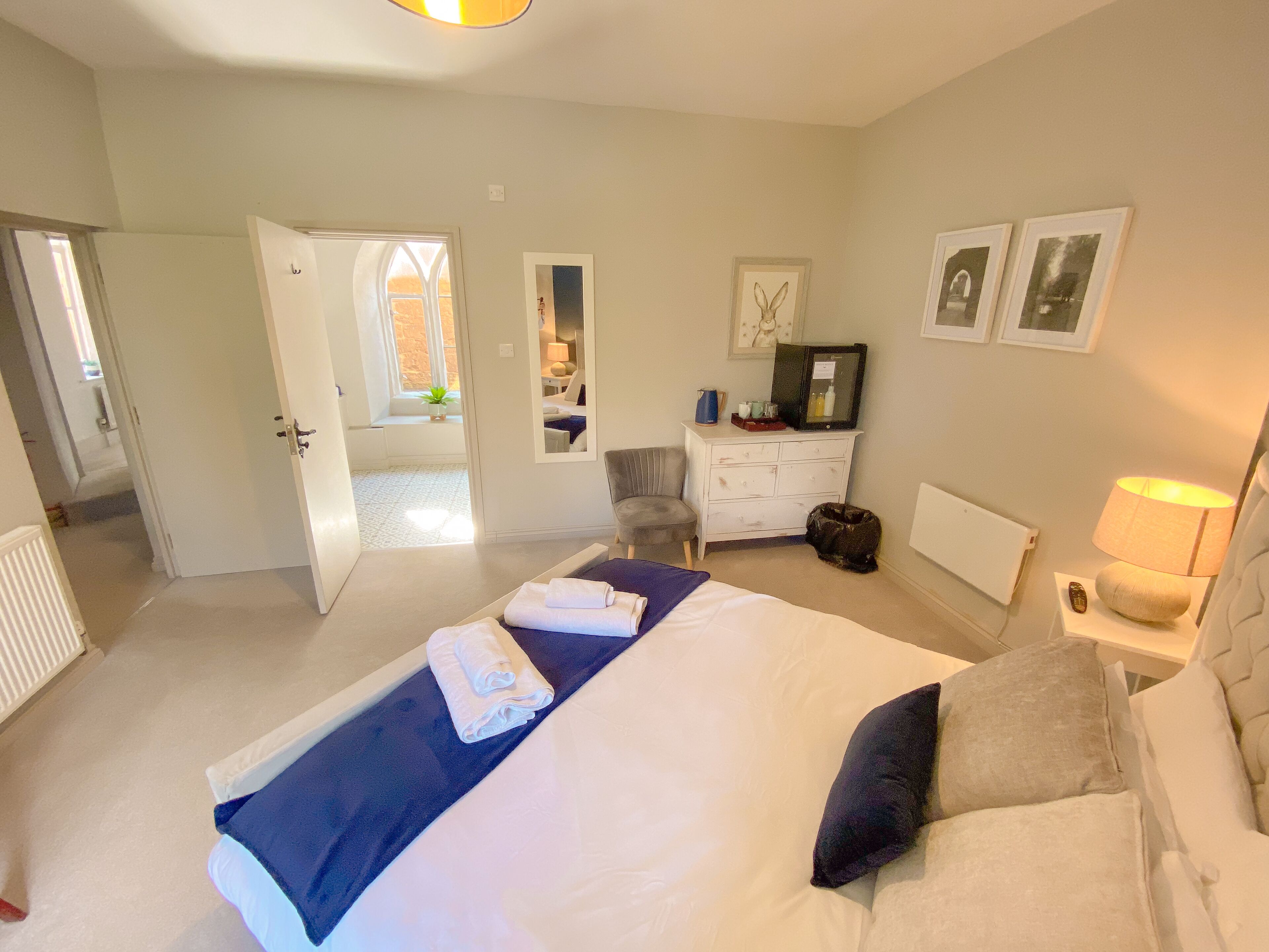 A bedroom with a large bed, a dresser, a chair, and a desk. There is a window and a door leading to another room.