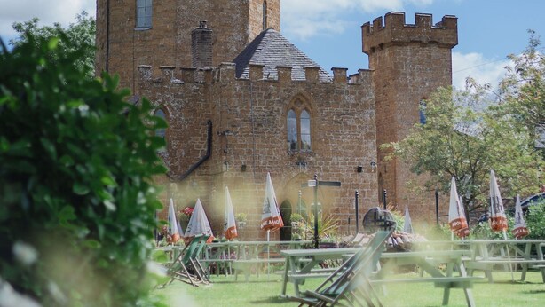 Un château historique doté d’une tour centrale et de deux tours plus petites, entouré d’une zone verdoyante avec des tables de pique-nique et des parasols.