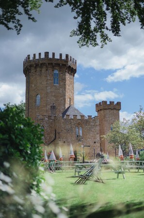 A historic castle with a central tower and two smaller towers, surrounded by a grassy area with picnic tables and umbrellas. - Castle at Edgehill (Banbury)