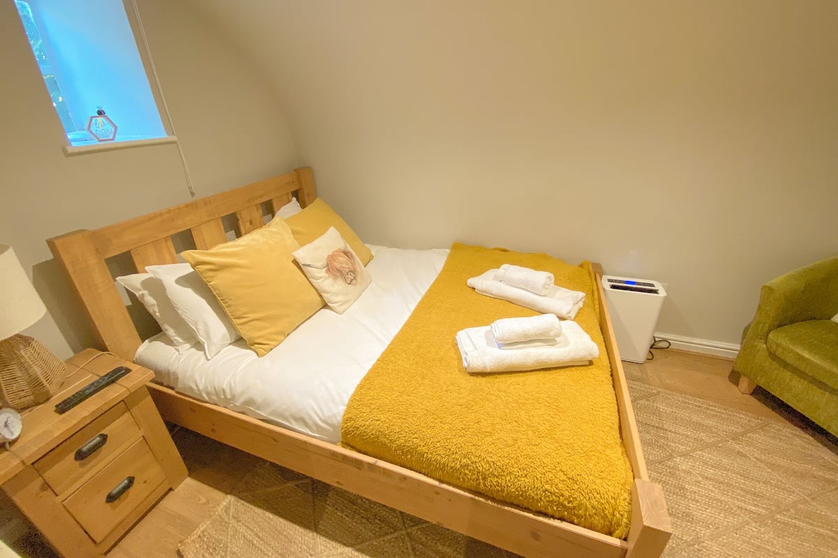 Cama bem arrumada com um cobertor amarelo, cabeceira de madeira e almofada com padrão floral. Um criado-mudo com abajur e relógio. Uma cadeira verde visível no canto.
