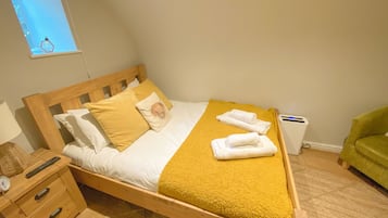 Una cama bien tendida con una colcha amarilla, cabecera de madera y un cojín con diseño floral. Se ve una mesita de noche con una lámpara y un reloj. Una silla verde es visible en la esquina.