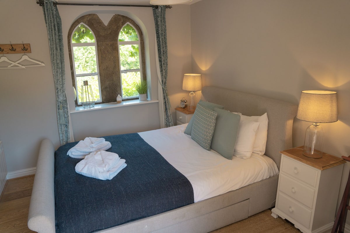 Cama bem arrumada com manta azul escura e almofadas brancas, mesa de cabeceira com abajur, e janela com cortinas.