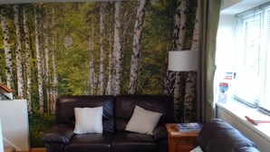A living room with a brown leather sofa, two patterned pillows, a small wooden side table with a book, and a window with blinds. - Bancroft Apartment (Durham)