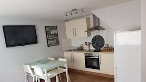 A compact kitchen with white cabinets, a black oven, and a white refrigerator. There is a dining table with white chairs and a black ottoman. A flat-screen TV is mounted on the wall, and a motivational quote is displayed above the counter. - Bancroft Apartment (Durham)