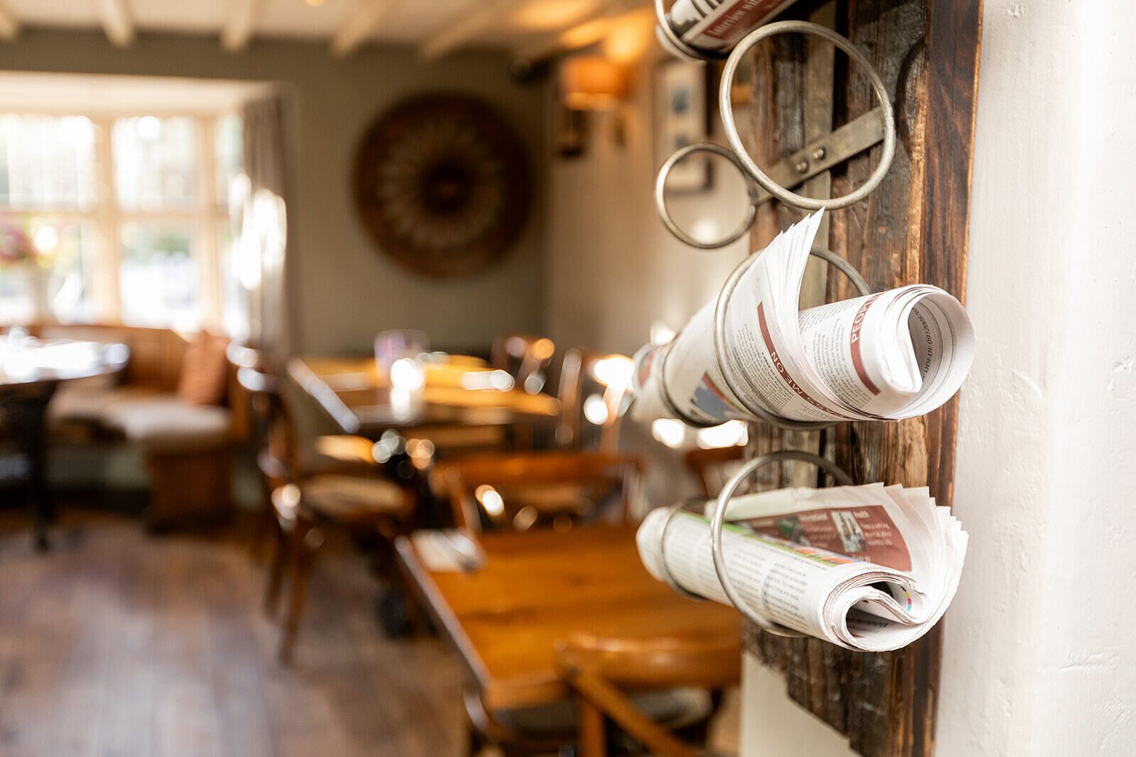 A restaurant interior with wooden tables and chairs, a wall-mounted newspaper rack, and a large decorative plate.