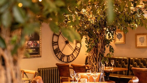 A restaurant interior with leather chairs, wooden tables, a large clock on the wall, and string lights. - The Dragon (Derby)