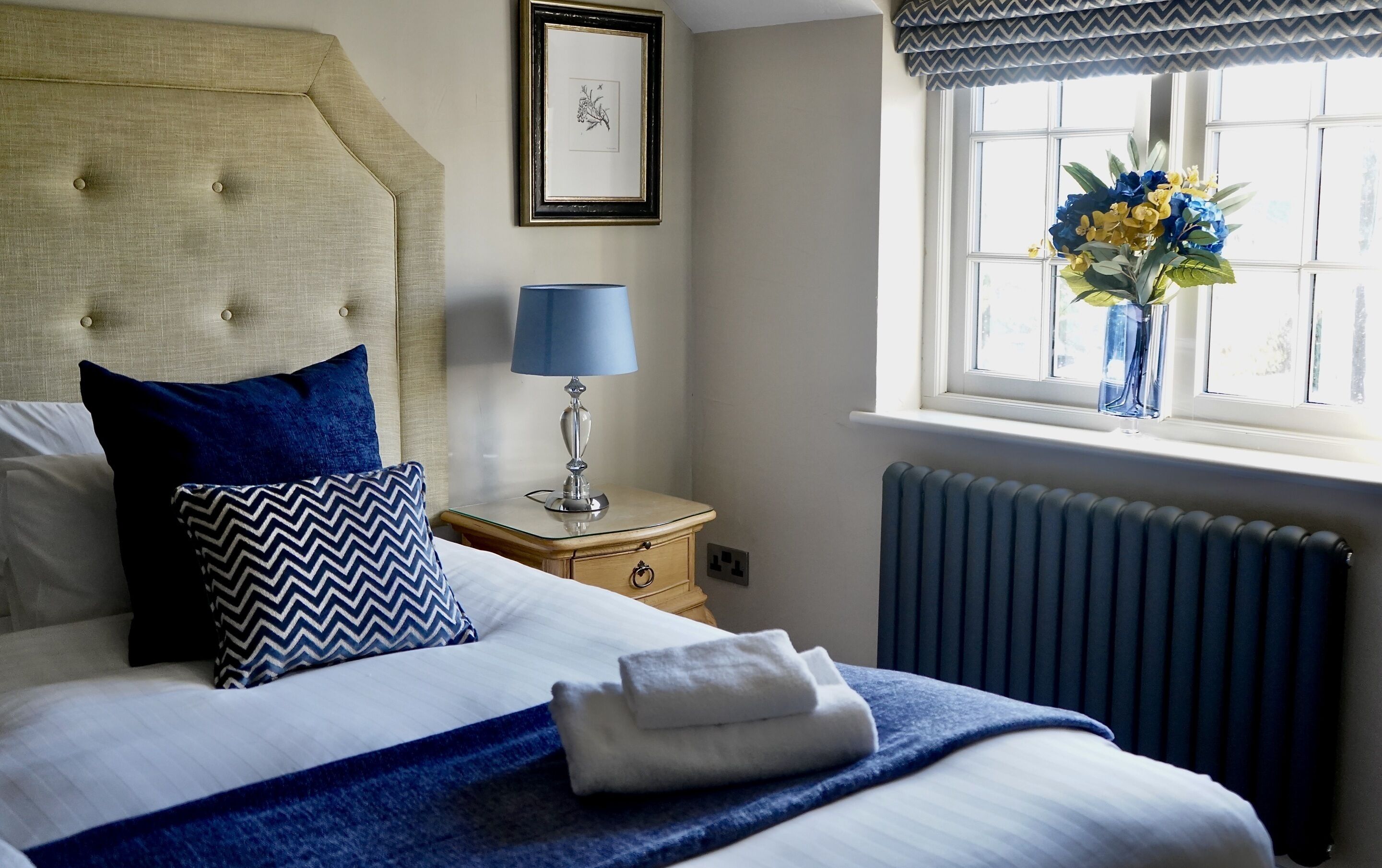 A neatly made bed with blue and white bedding, a bedside lamp, a framed picture on the wall, and a window with a view of flowers.