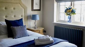 A neatly made bed with blue and white bedding, a bedside lamp, a framed picture on the wall, and a window with a view of flowers.