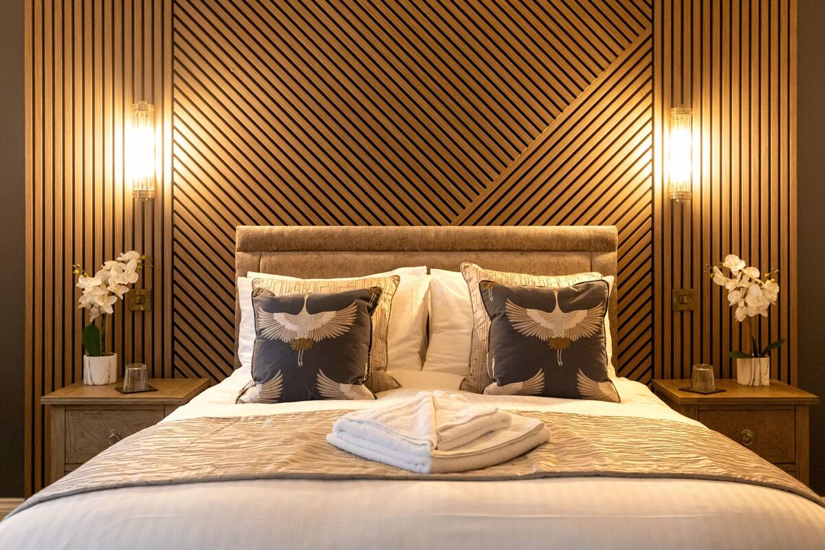 Una cama bien hecha con una colcha estampada, dos almohadas con diseño de pájaros y dos mesitas de noche con lámparas y jarrones de flores.