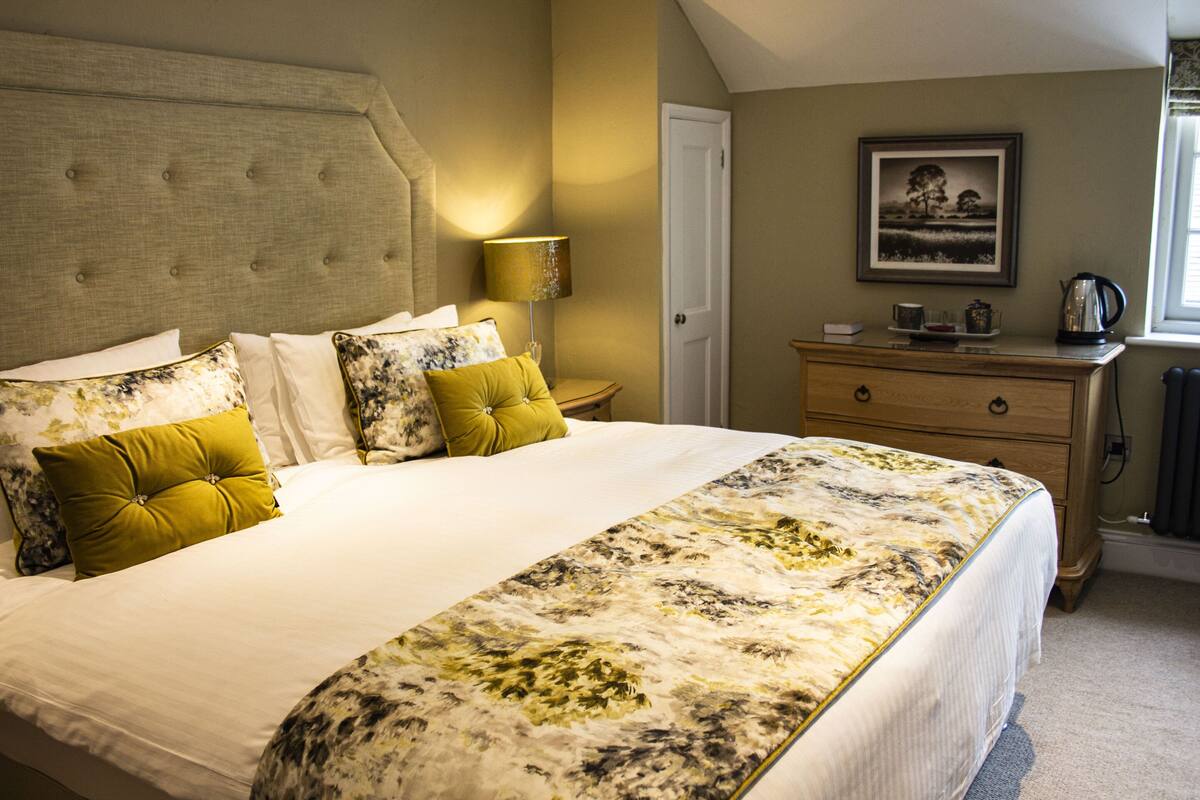 Una cama bien hecha con una colcha estampada con motivos florales, una lámpara de noche y un aparador con un cuadro enmarcado encima.