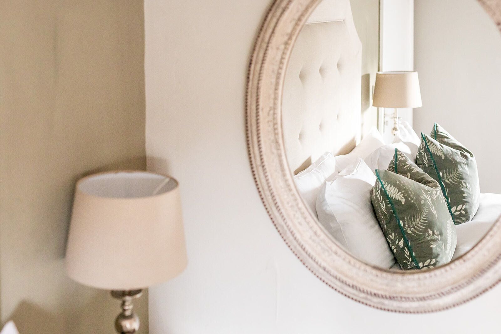 A bedroom with a round mirror, a bedside lamp, and decorative pillows.