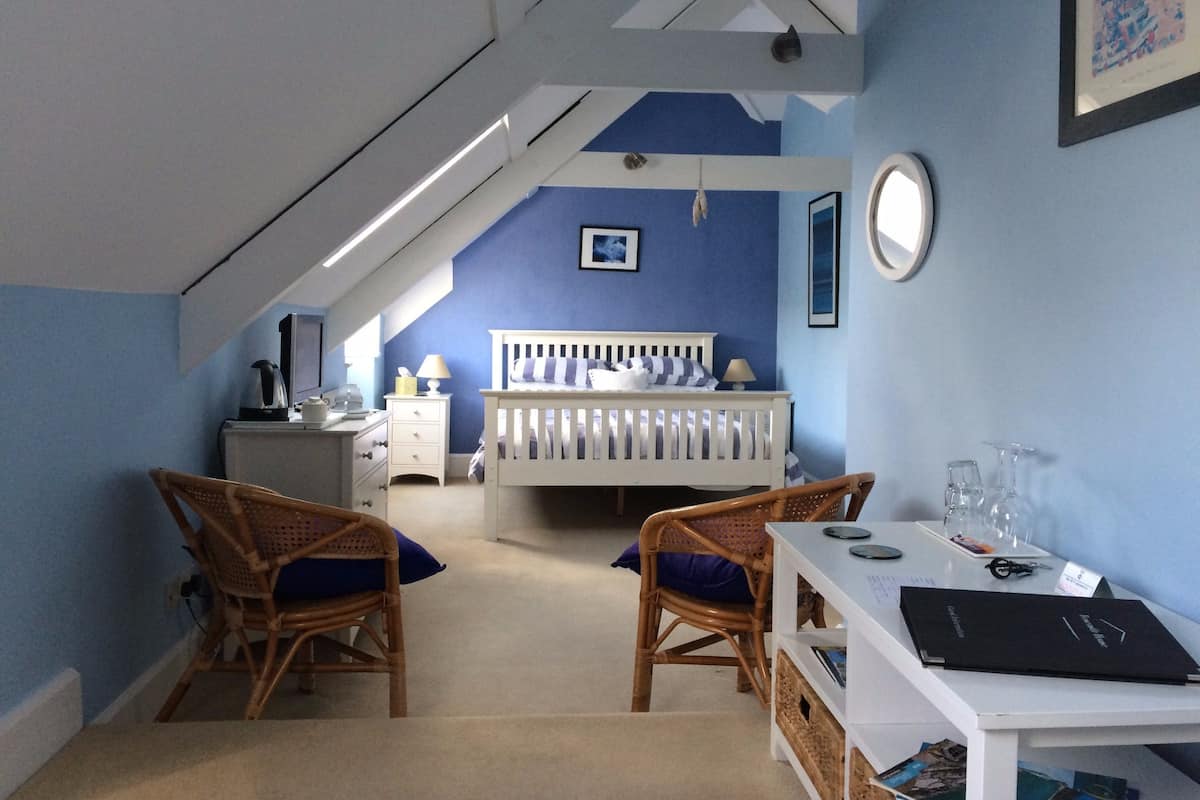 A bedroom with a sloped ceiling, a white bed, two wicker chairs, a desk with a computer and books, and a small window.