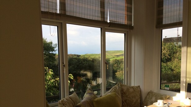 A room with a large window offering a view of green fields and trees. There is a wooden table with a cup and saucer, and a chair with cushions. - Boscastle House (Boscastle)