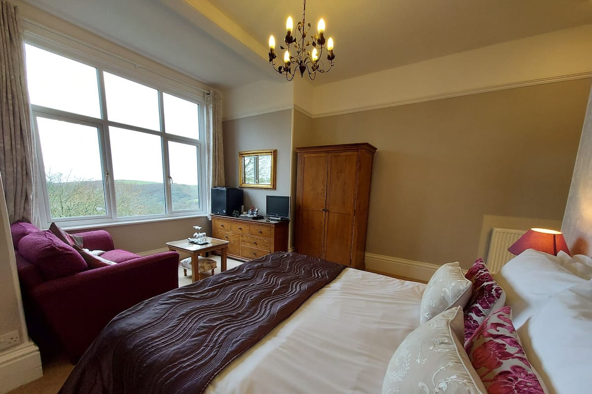 A hotel room with a large bed, a wooden wardrobe, a desk with a television, a chair, and a view of the outdoors through a large window.