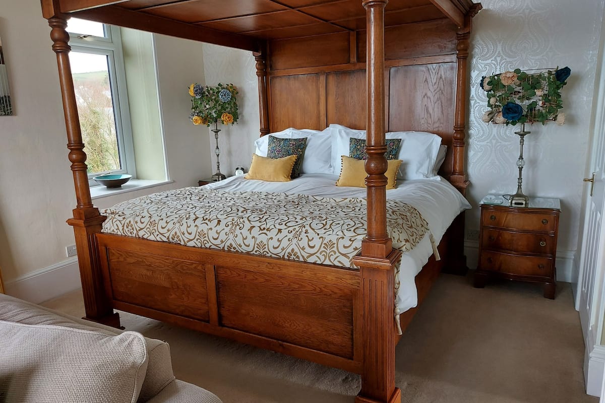 A four-poster bed with a patterned bedspread, a bedside table with a lamp, and a window with a view of the outdoors.