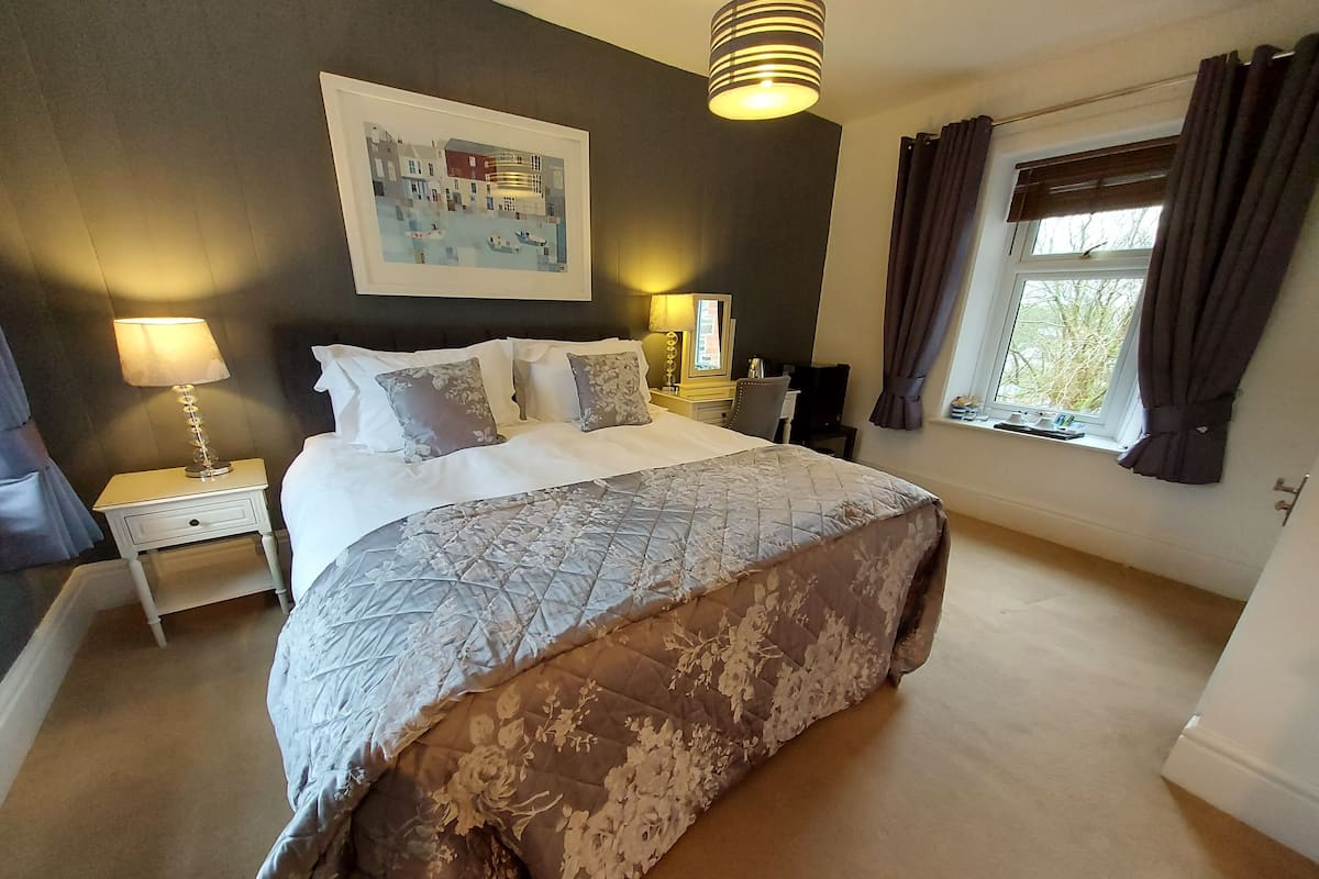A bedroom with a large bed, a bedside table with a lamp, a framed picture on the wall, and a window with curtains.