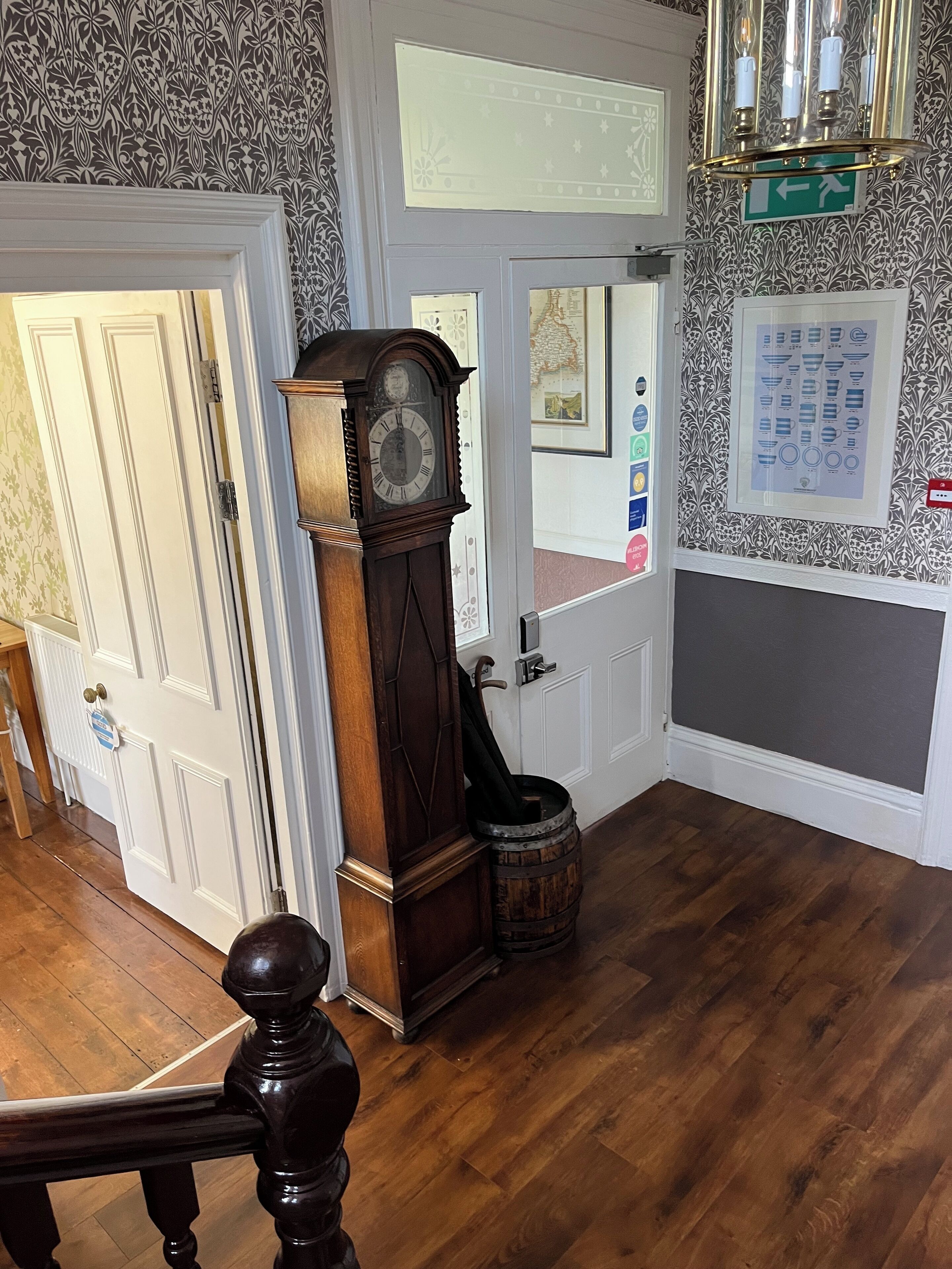A room with a wooden floor, a grandfather clock, a door with a sign, and a wall with a framed diagram.