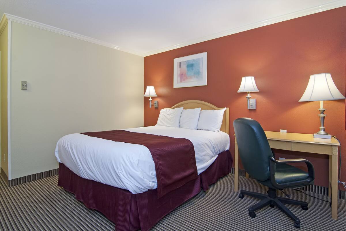 Una habitación de hotel con cama, escritorio, silla y dos lámparas fijadas a la pared.