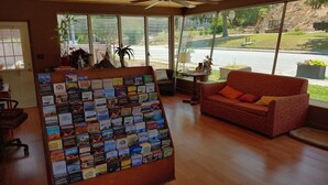 A room with a wooden floor, a red patterned sofa, a display of books, and a view of a construction site through large windows. - RoseWood Inn (Bryson City)