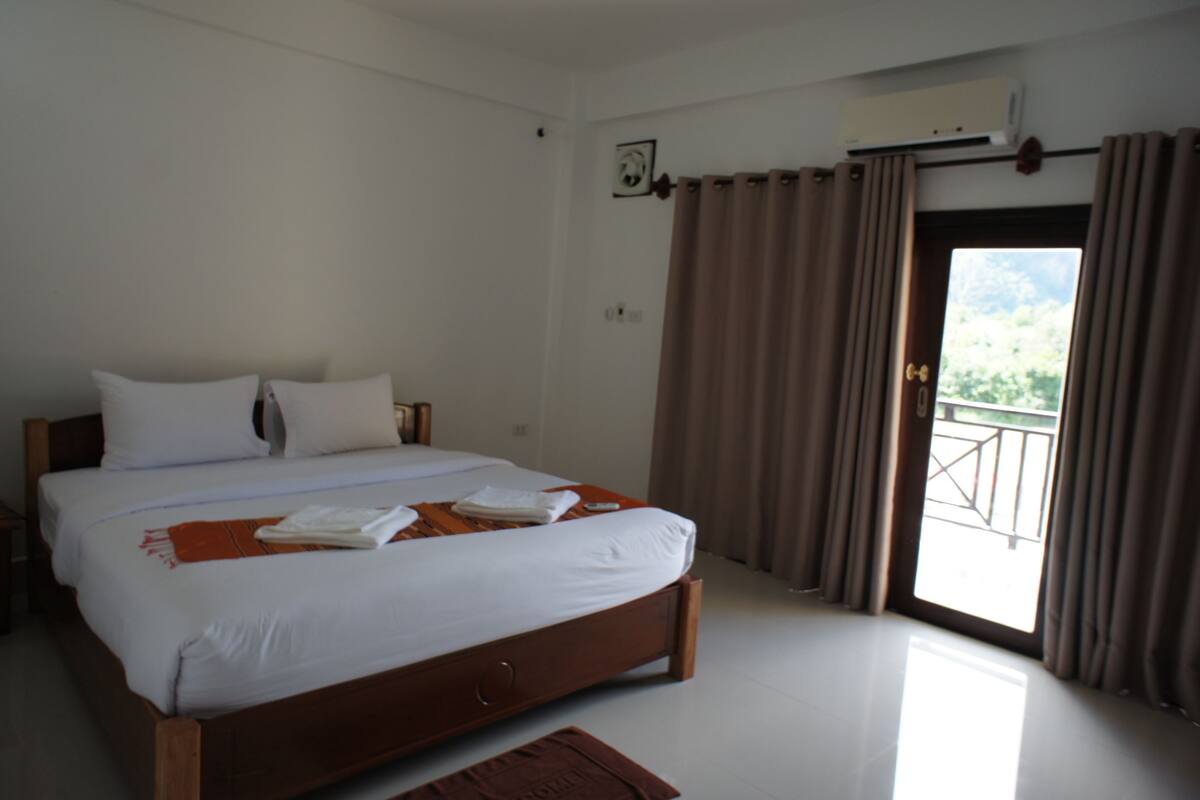 A neatly made bed with white linens and a brown headboard, a wooden nightstand, and a view of a balcony through a door with curtains.