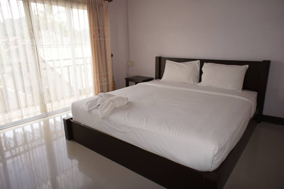 A neatly made bed with white linens and a dark headboard in a room with a large window and a small bedside table.