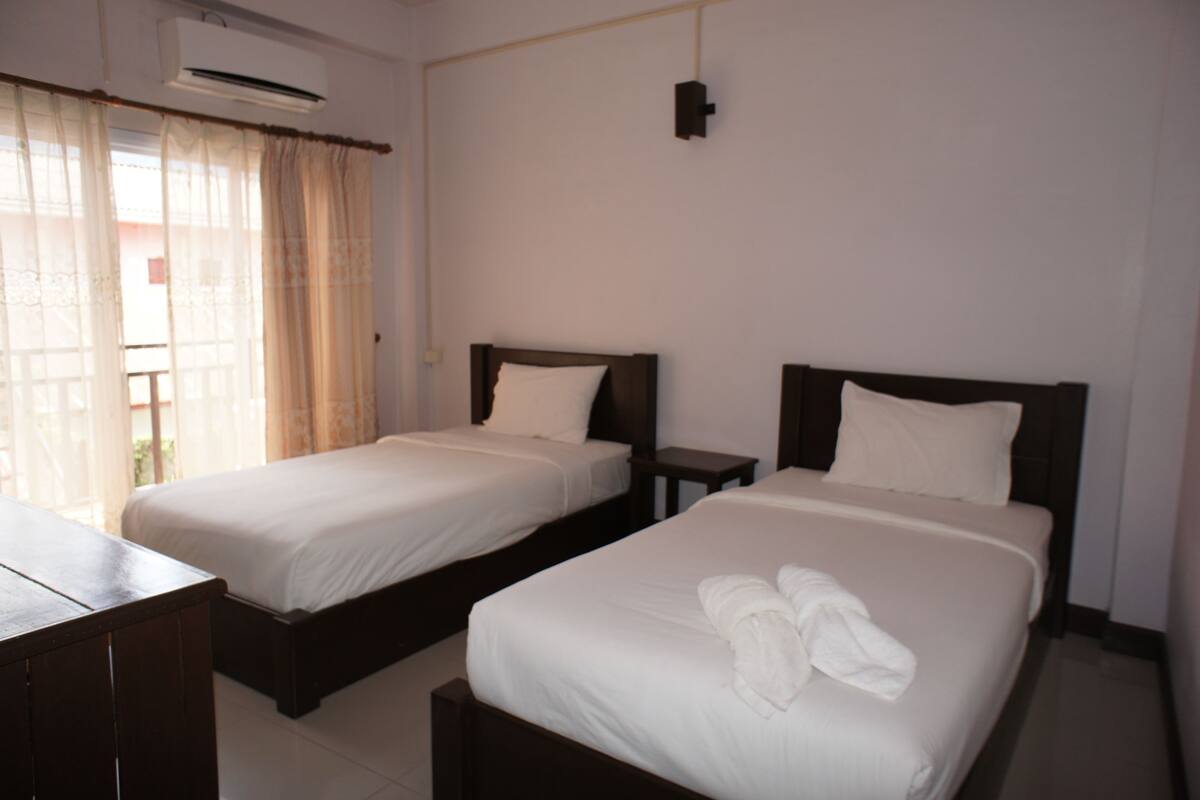 A hotel room with two single beds, a wooden desk, and a window with sheer curtains.