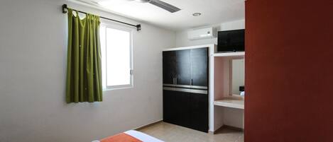 Habitación de hotel con cama, ventilador de techo, ventana con cortinas verdes, armario negro y una pared de acento roja.