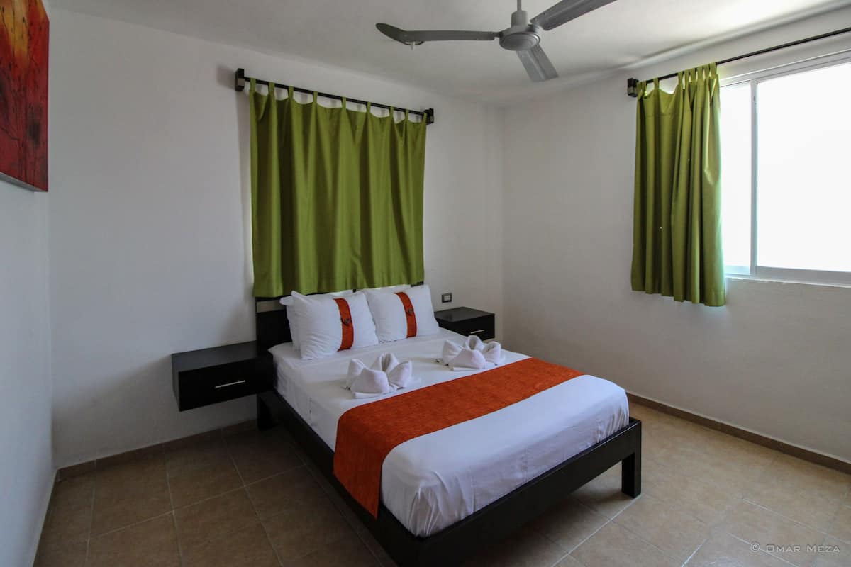 A neatly made bed with white linens and an orange accent, a black nightstand, a ceiling fan, and green curtains.