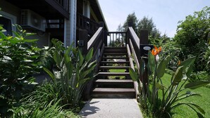 En trætrap fører op til en bygning med balkon, omgivet af frodig vegetation og en velplejet græsplæne.