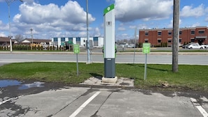 Un espace de stationnement équipé de bornes de recharge pour véhicules électriques, un panneau vert indiquant une place de stationnement réservée, et un bâtiment en briques en arrière-plan.