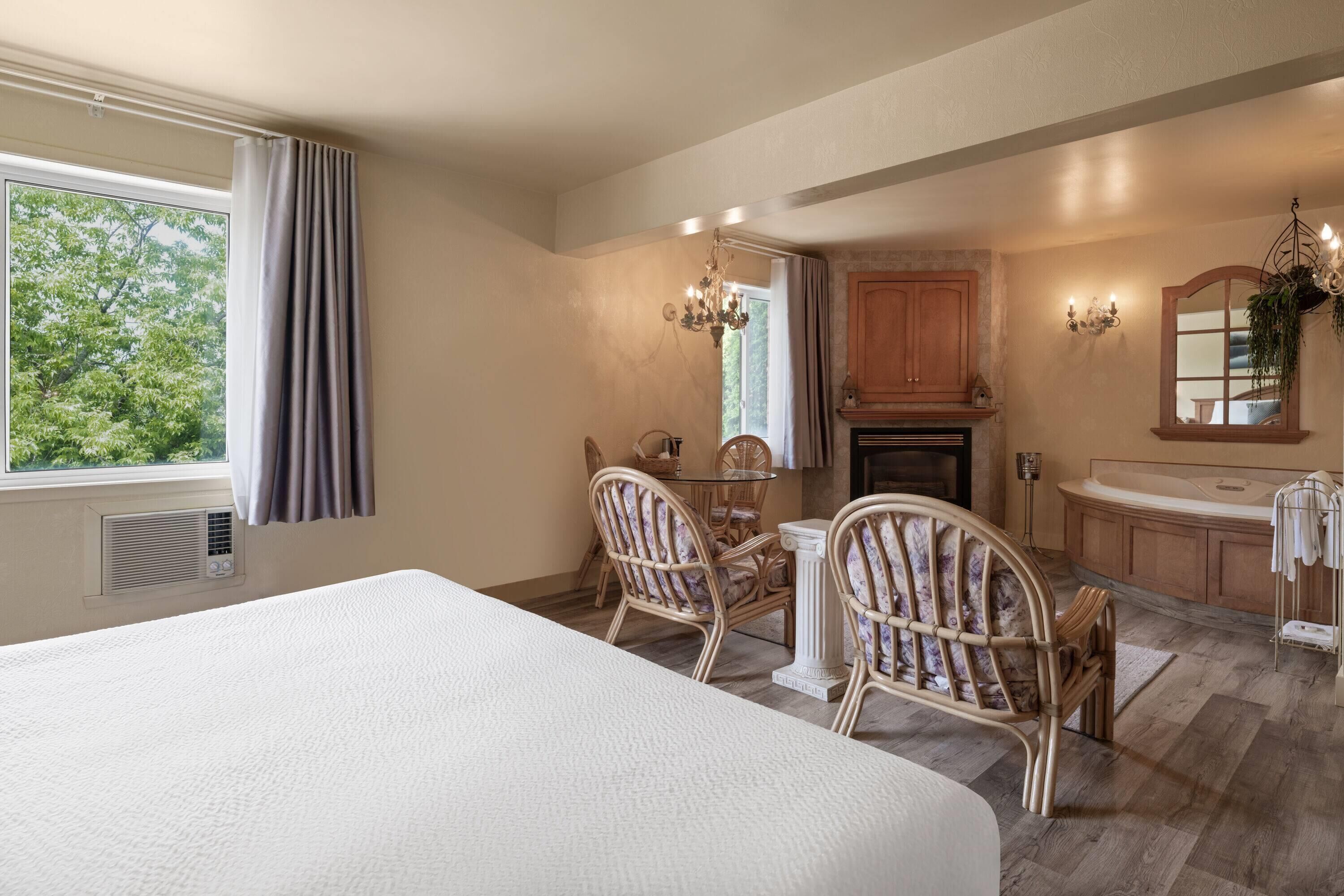 Habitación de hotel con una cama grande, dos sillones de mimbre, una mesita, una chimenea, un espejo y una ventana con cortinas.