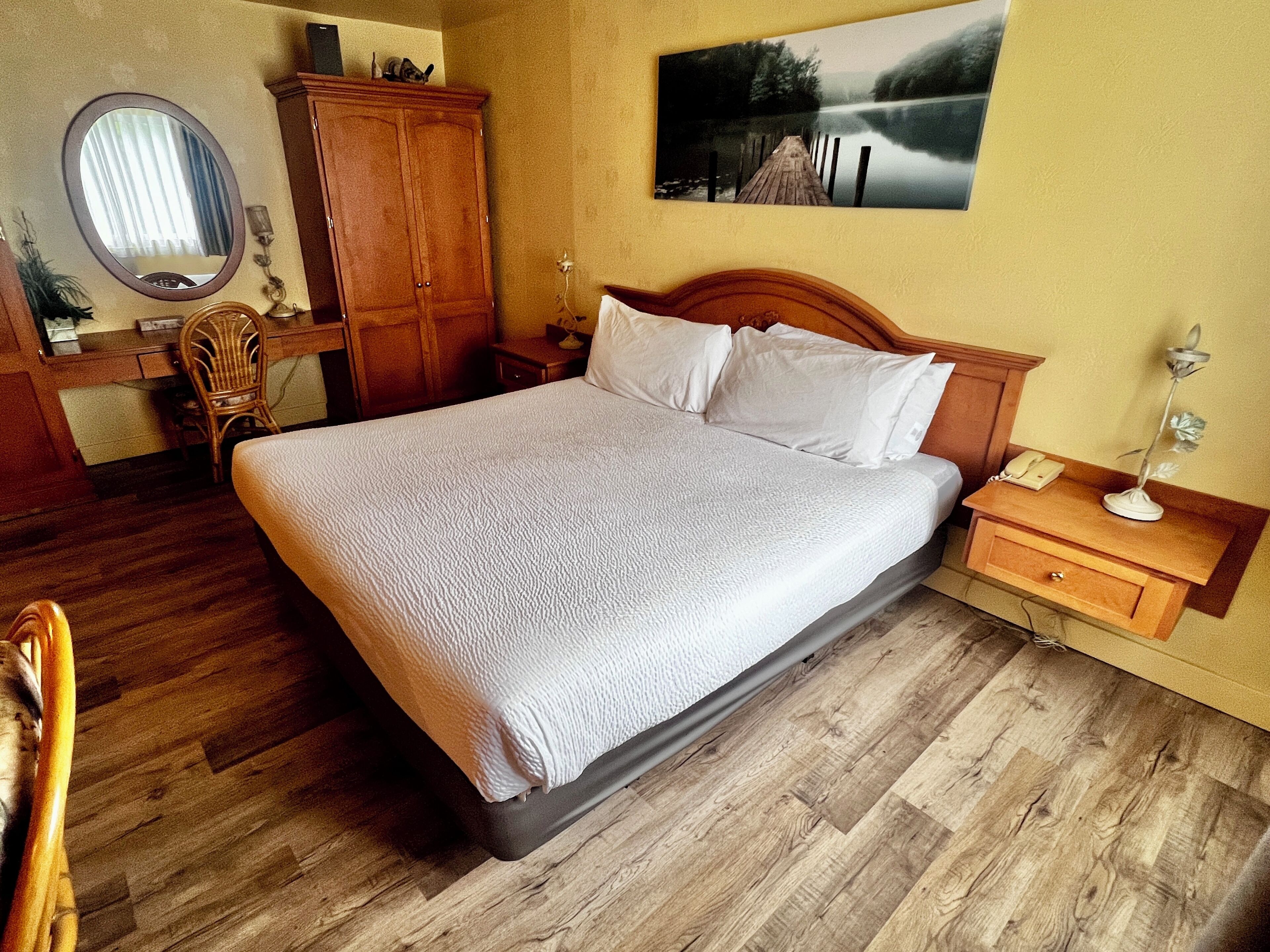 Een keurig opgemaakte slaapkamer met een groot bed, een houten garderobe, een bureau met stoel en een nachtkastje met lamp.
