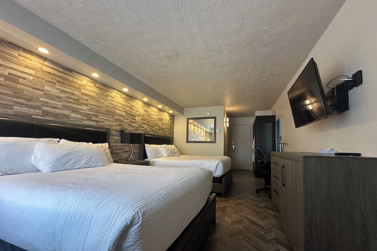 Quarto de hotel com duas camas, televisão na parede, cabeceira de madeira e chão com padrão herringbone.