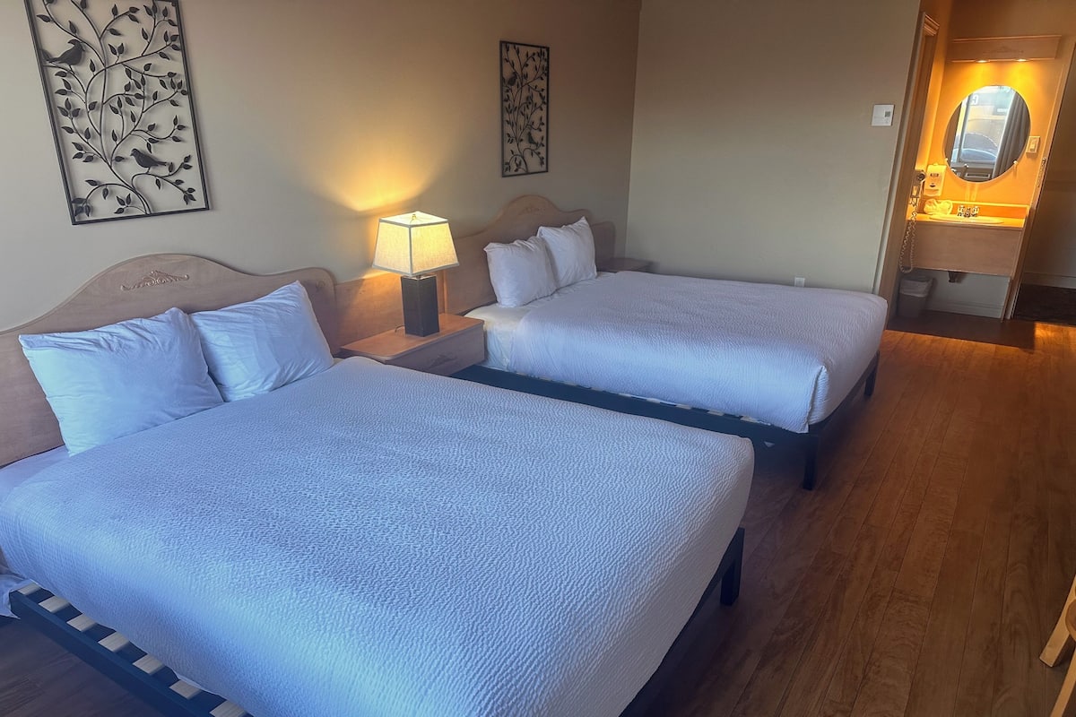 Quarto de hotel com duas camas, uma mesa de cabeceira com abajur e um banheiro visível por uma porta aberta.
