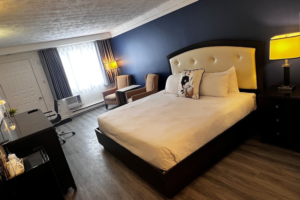 Quarto de hotel com uma cama grande, uma escrivaninha, uma cadeira, um abajur e vista para o exterior.