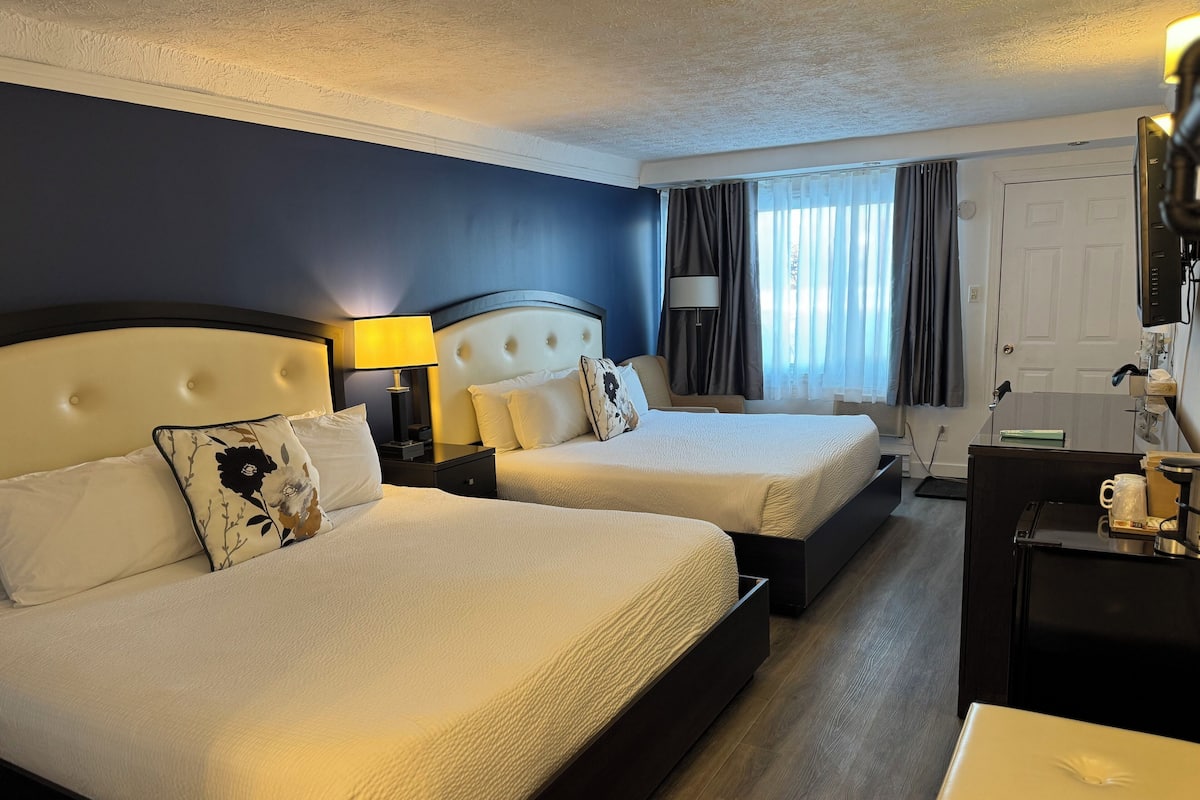 Quarto de hotel com duas camas, uma escrivaninha, uma cadeira, um abajur e uma janela com cortinas.