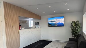 Un espace d’accueil avec un comptoir de réception en marbre, un paillasson noir portant l’inscription « Travelodge », un téléviseur mural diffusant un paysage pittoresque et une plante en pot.