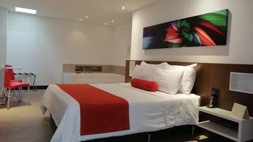 A modern hotel room with a large bed, a desk with a chair, a small kitchenette, and a large painting on the wall.