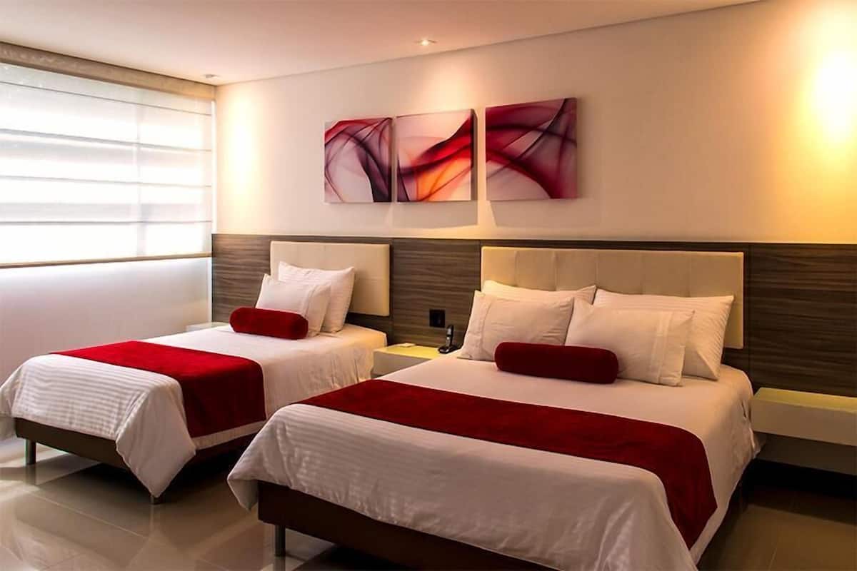 A hotel room with two beds, each with a red accent pillow, white bedding, and a modern headboard. There are abstract wall art pieces above the beds and a nightstand with a lamp between them.