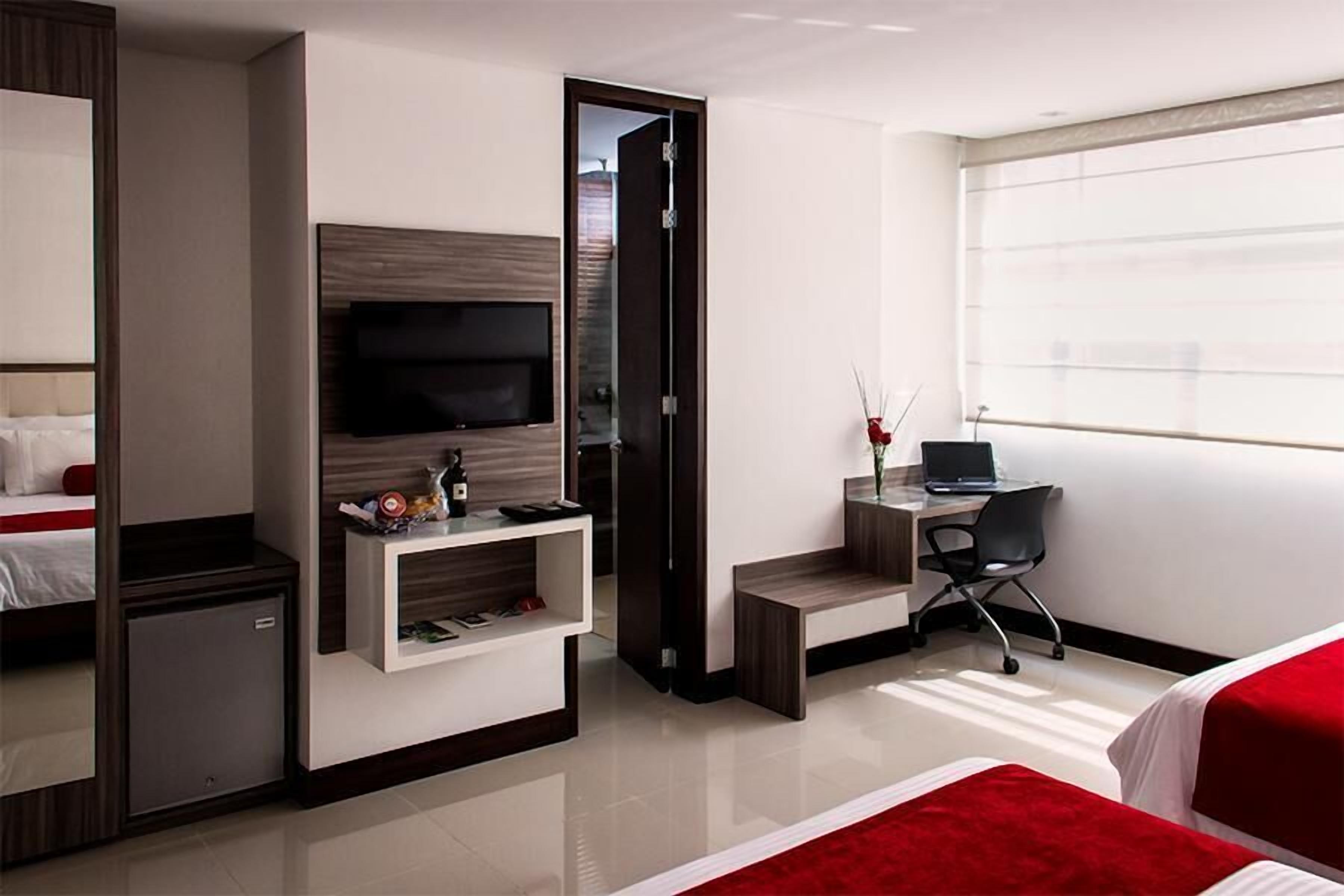 Una habitación de hotel con una cama, un escritorio con una computadora portátil, un televisor y un refrigerador pequeño.