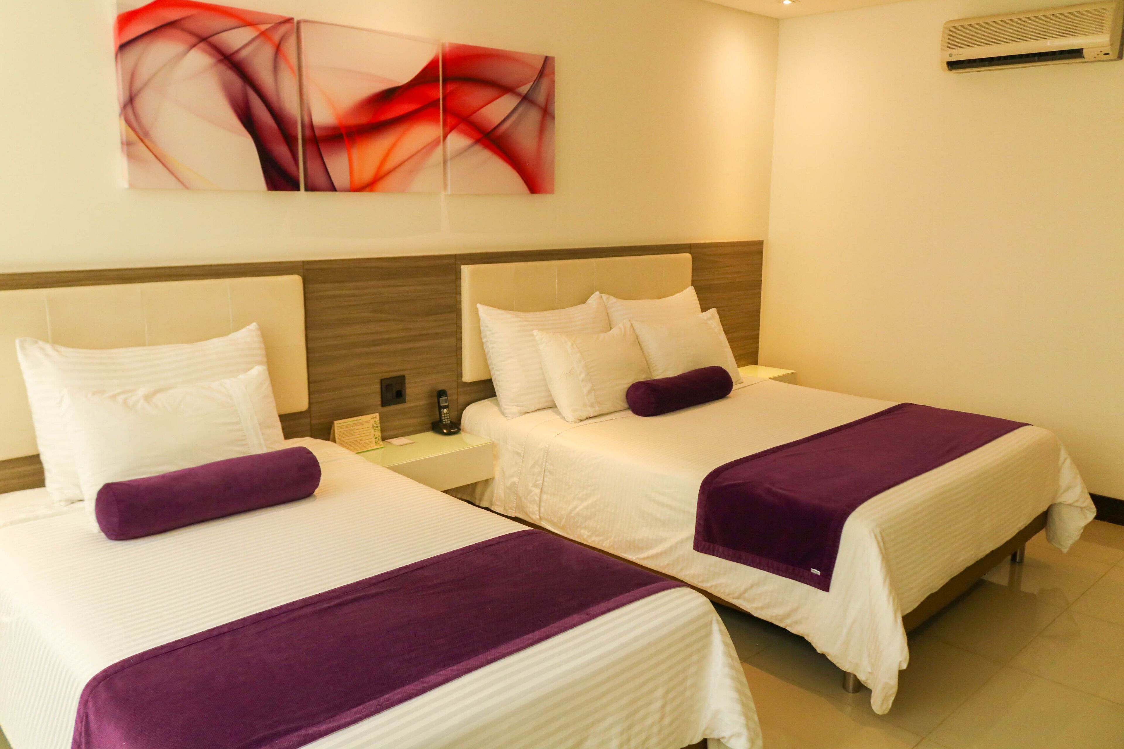Habitación de hotel con dos camas, cada una con ropa blanca y detalles morados, un cuadro abstracto moderno en la pared y un equipo de aire acondicionado.
