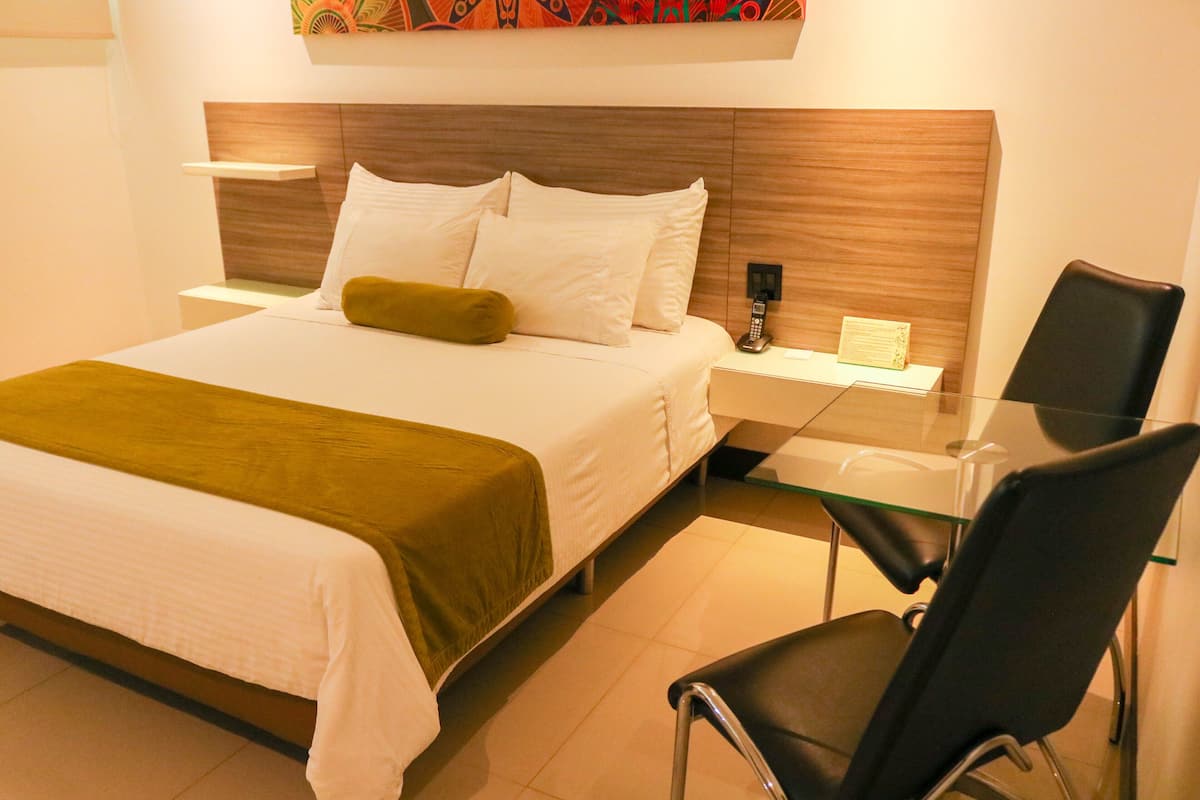 A hotel room with a large bed, a bedside table with a lamp, a chair, and a small desk with a telephone.