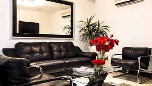 Una sala de estar moderna con un sofá de cuero negro, una mesa de centro de vidrio y un sillón de cuero negro. Hay un espejo grande en la pared y una planta en maceta con flores rojas.