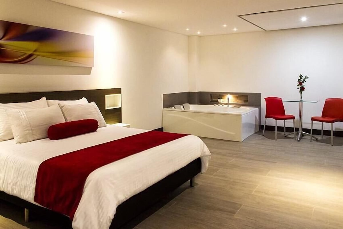 A modern hotel room with a large bed, a small desk, and a seating area with red chairs.